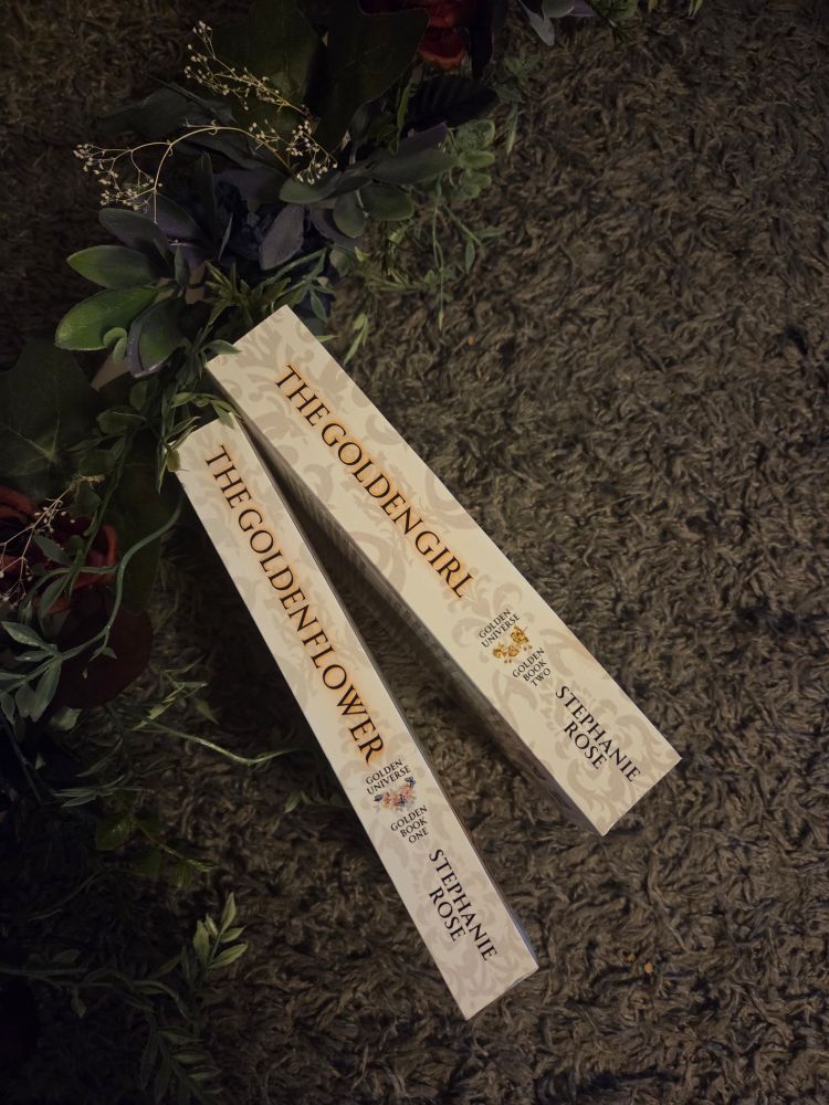 Picture of the spines of two books - The Golden Flower & The Golden Girl, by Stephanie Rose - with dark flowers and leaves overhead