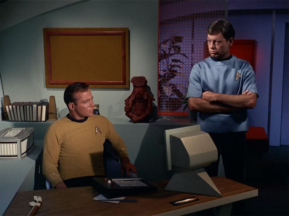 Kirk at his desk, with McCoy beside him. 