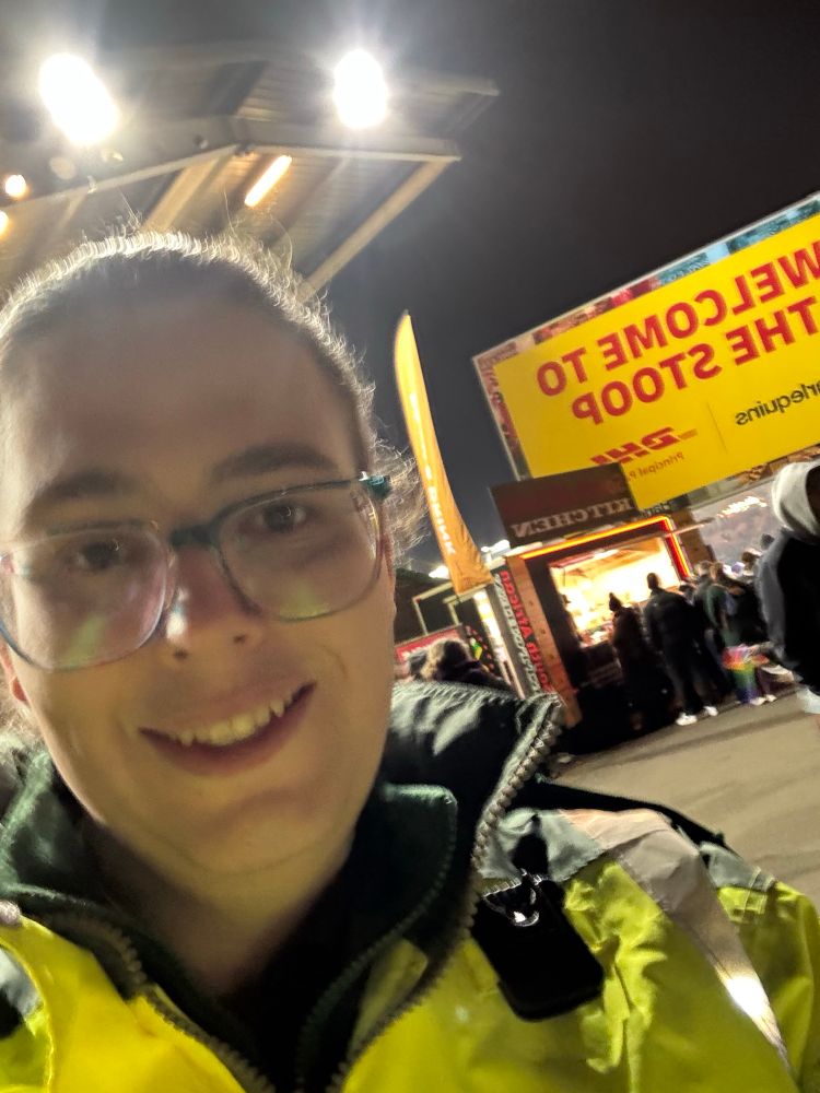 Chloe standing in front of a “Welcome to the Stoop” sign at a rugby stadium wearing ambulance uniform with a high vis jacket on. 