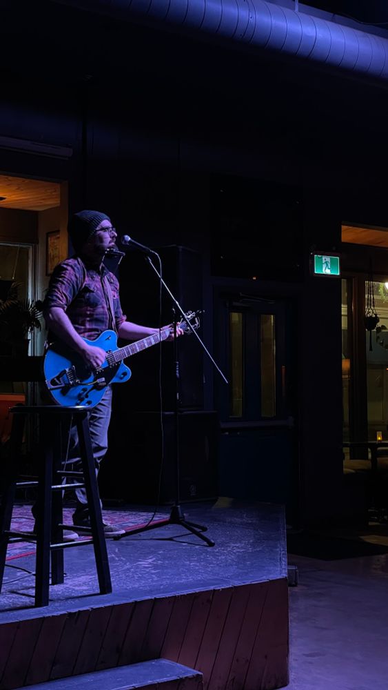 Jeremy stands on stage playing guitar. There is a kazoo in a harmonica harness around his neck. 