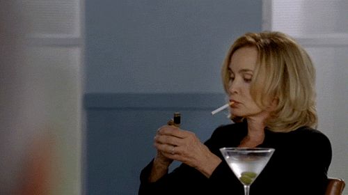 A GIF of Jessica Lange in her role as Fiona Goode in the television show American Horror Story: Coven. She is sitting down with a martini in front of her while she lights a cigarette. 