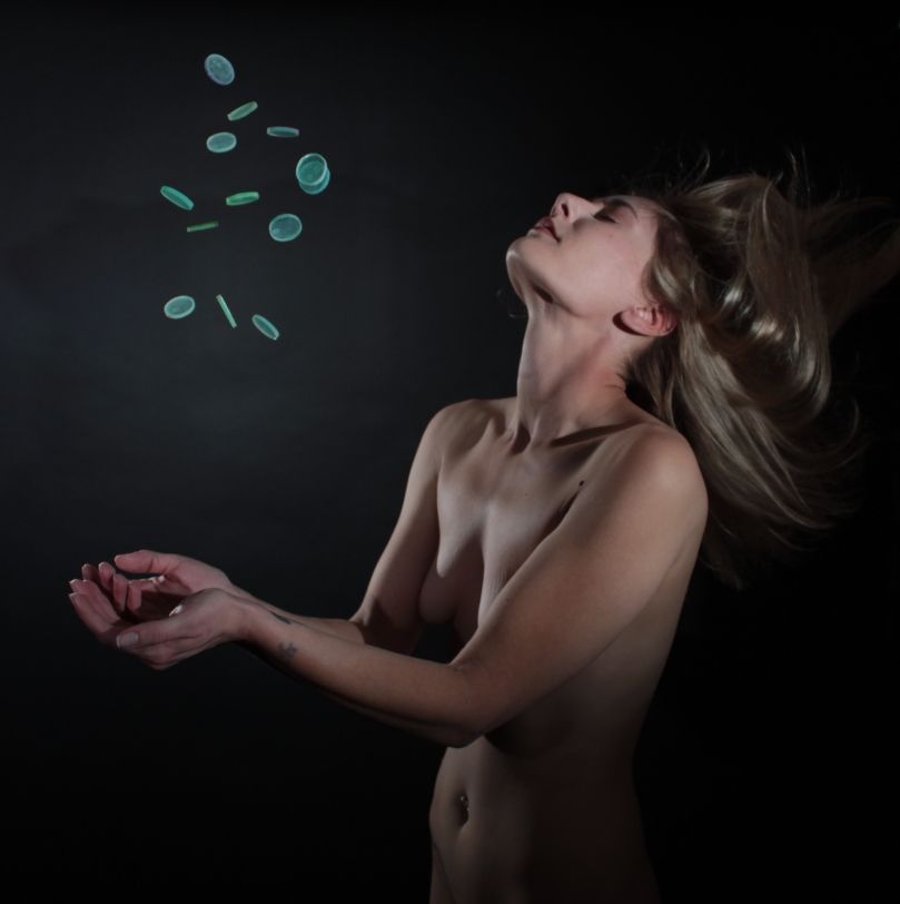 Nude art model tosses green disks in the air.  Studio shot with black background.