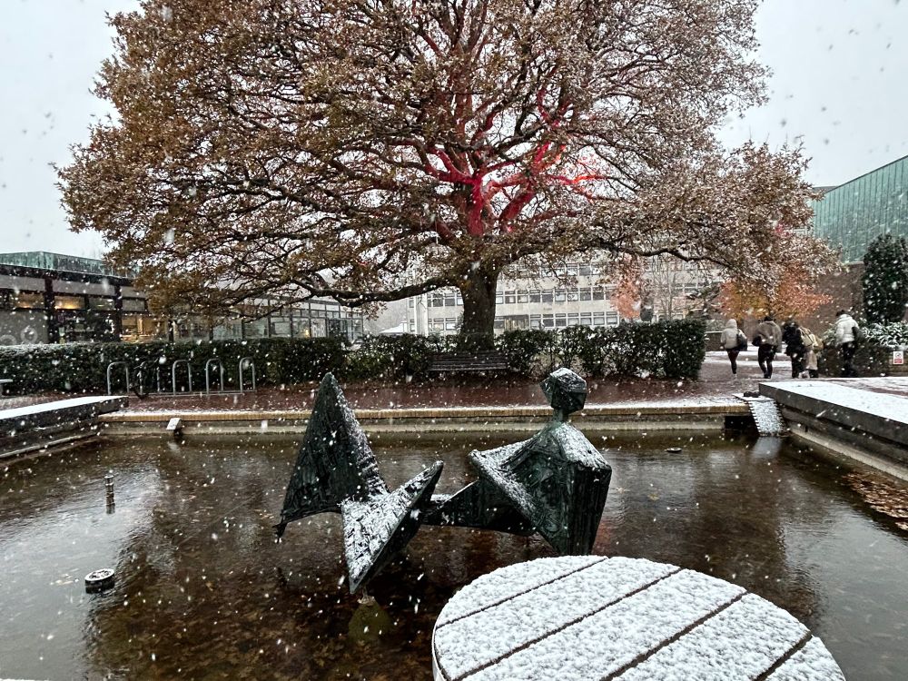 Mc Williams Puy de Dôme sculpture on water with snow coming down with oak tree behind & snow on round picnic table front right 