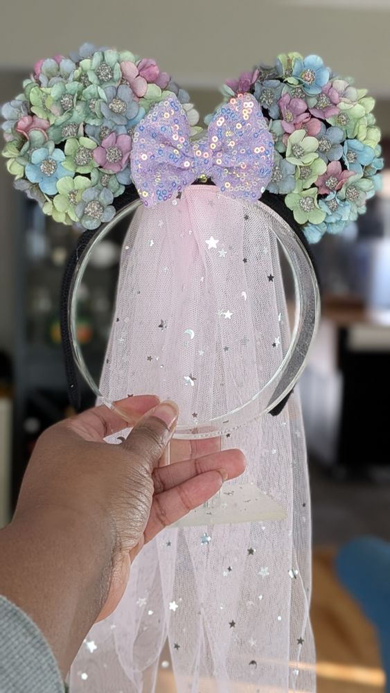 Marcy holds another version of the floral ear headband featuring mint green, lavender, and baby blue flowers with a lavender sequin bow at the center. A soft pink tulle veil decorated with tiny metallic moons and stars flows down behind the headband