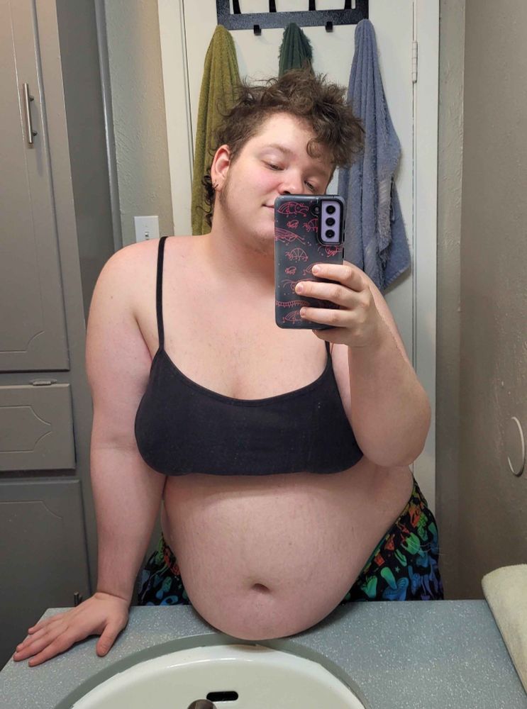 Mirror selfie of me, a fat pale skinned person with a curly brown mohawk, standing against the sink, my tummy resting on the counter. 