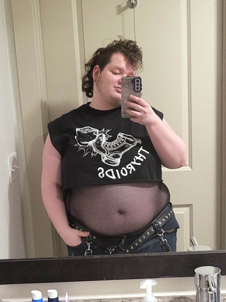 Mirror of a fat pale skinned person with a curly brown mohawk. He's wearing a black cropped sleeveless Thyroids band shirt, a mesh tank under it, and jeans with the belt undone, his tummy hanging over it.
