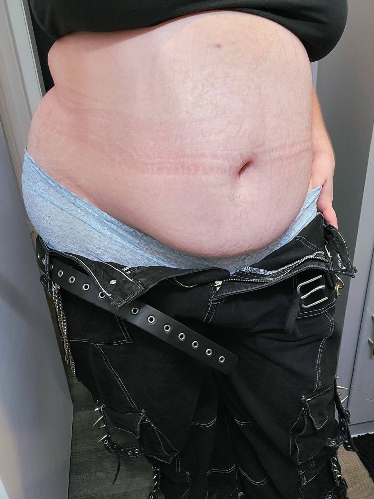 A fat pale skinned person with their pants pulled down to their thighs, showing off their tummy with indents from the waistband and lacy blue underwear. Their pants are big and black with an undone belt, spikes, chains, and many pockets. Their thumb is teased under the waist of the underwear.