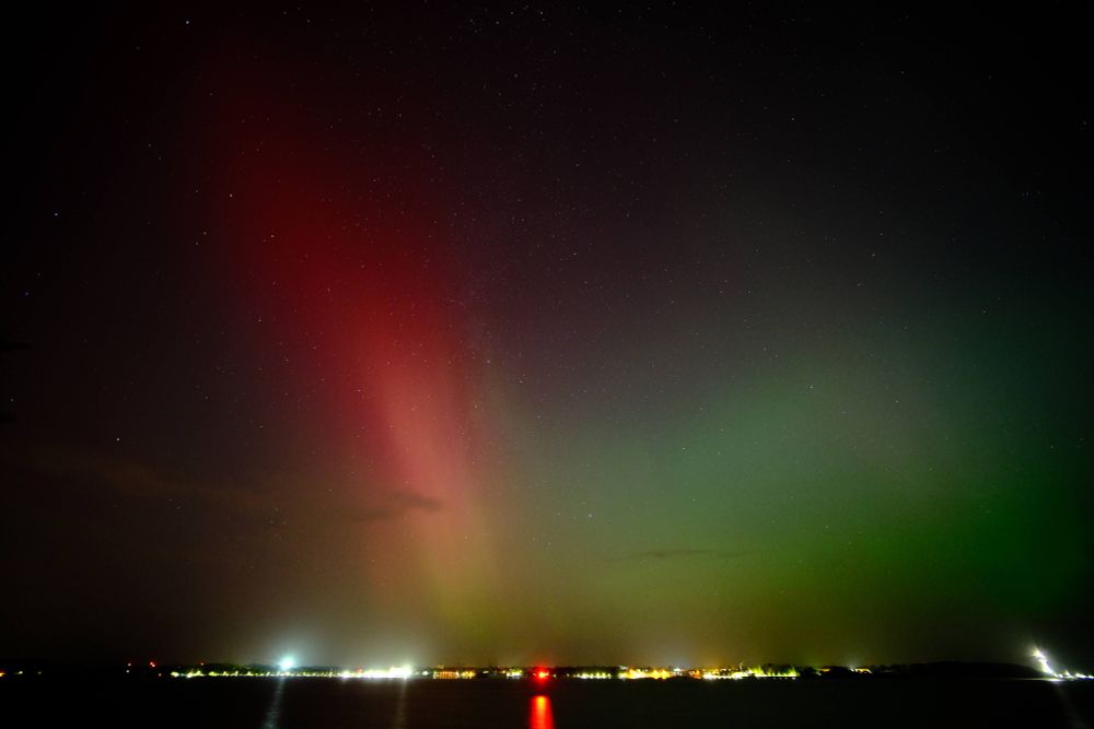 Rotes und grünes Polarlicht mit Sternenhimmel dahinter. Am unteren Bildrand ein paar Lichter.