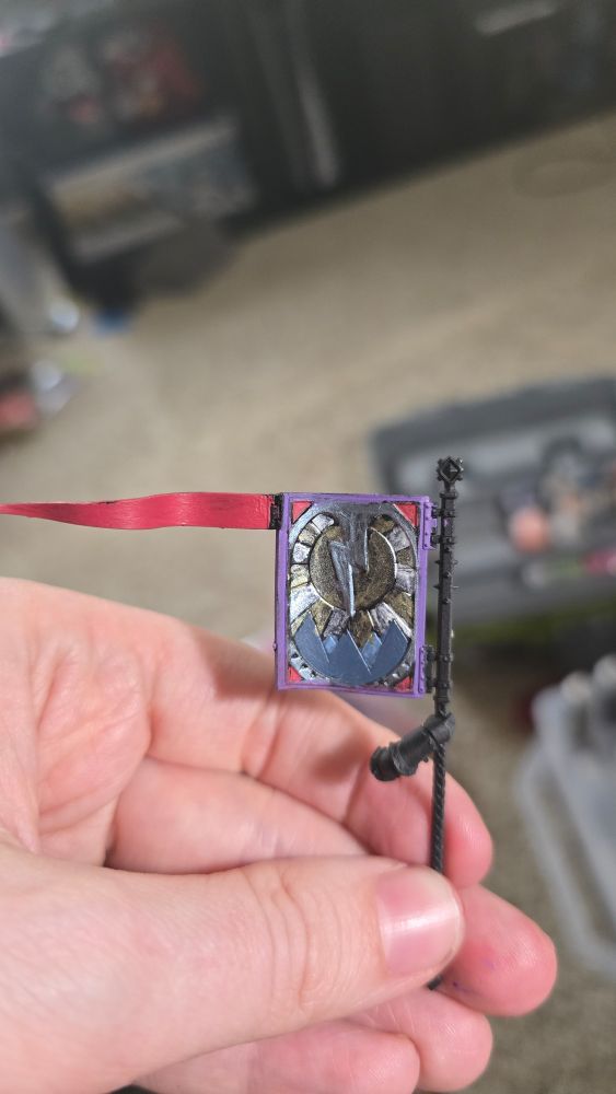 A white hand holds a plastic Age of Sigmar Stormcast Eternal flag and flag pole. The flag has a lighting bolt painted silver striking over blue mountains as rays of sunlight break behind the bolt. The flags boarder is painted light purple with a red tail flag.