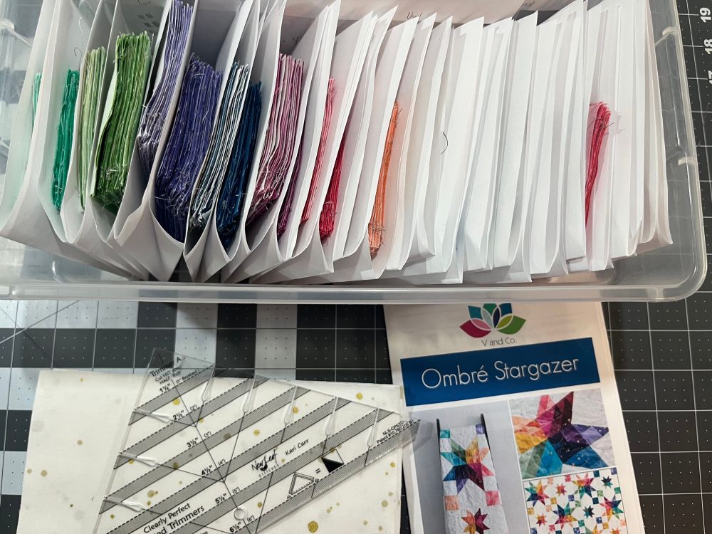 A shoebox filled with many colors of fabric separated by pieces of paper sits on a cutting mat. Next to the box is a quilt pattern titled V. and Co Ombré Stargazer. A ruler designed to help trim half square triangles sits on a pile of larger pieces of white background fabric 