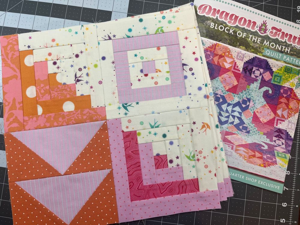 A stack of 4-patch quilt blocks consisting of 2 log cabin blocks, a concentric squares block, and flying geese block in shades of pink and orange sit on top of a pattern book titled Dragon Fruit Block of the Month 
