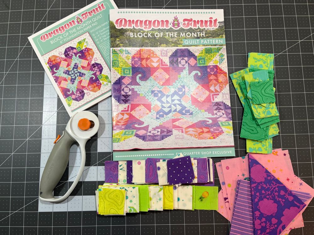 Cut fabrics in shades of green and purple are stacked on a cutting mat, surrounding a pattern book titled Dragon Fruit Block of the Month