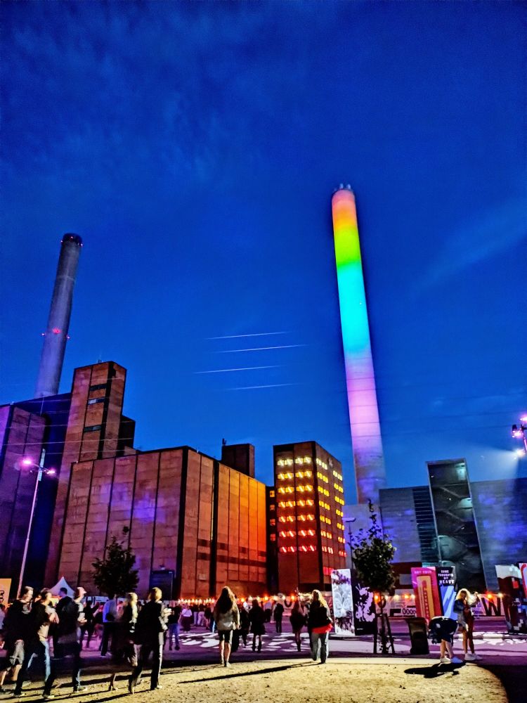 People walking at night towards a large lit-up reddish brown building with rainbow colors projected on a tall smokestack 