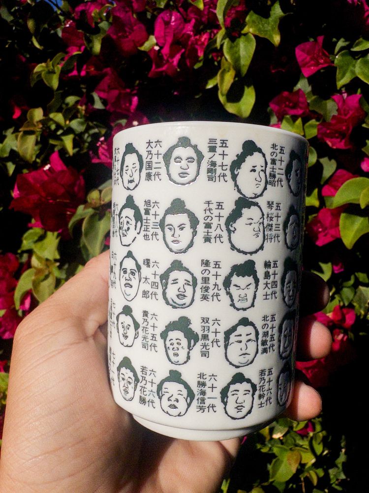 A light off white handle-less mug, with inset caricatures of the sumo champions from the past.