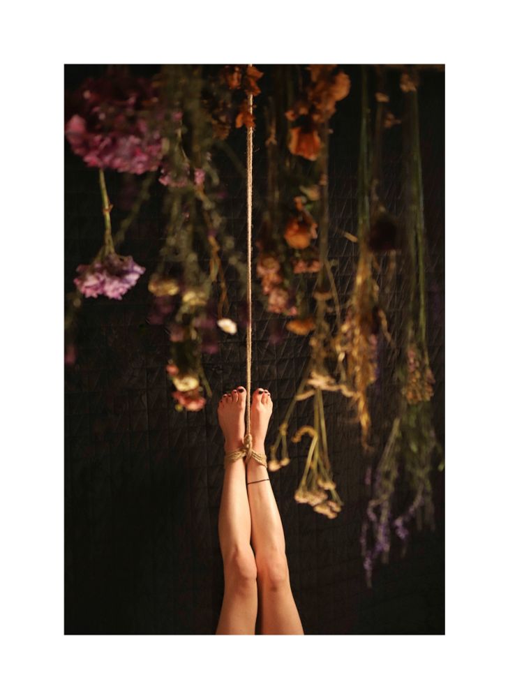 Image shows phot0 of dried flowers hanging from the ceiling. Also in background there is a pair of legs tied up and hanging tied by rope. To a black background.