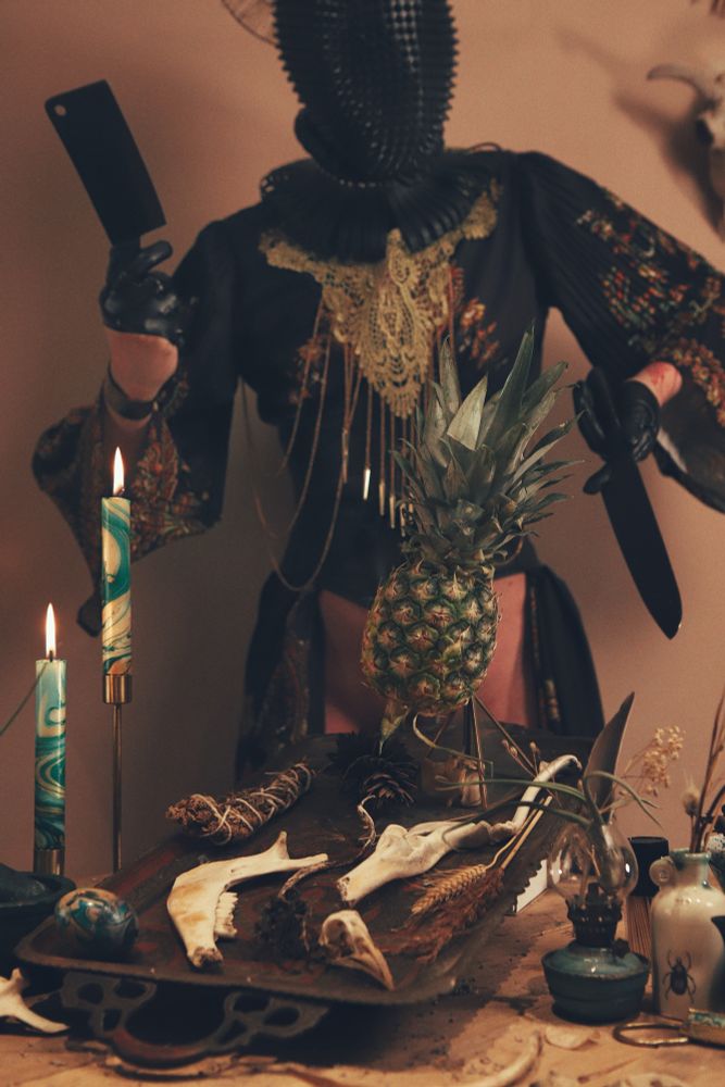Phot shows latex hooded figure in a gold green and black dress holding a knife and meat cleaver about to cut up a pineapple which is floating in front of them. A tray is also floating on a table in front of them which feature bones, skulls and painted eggs.