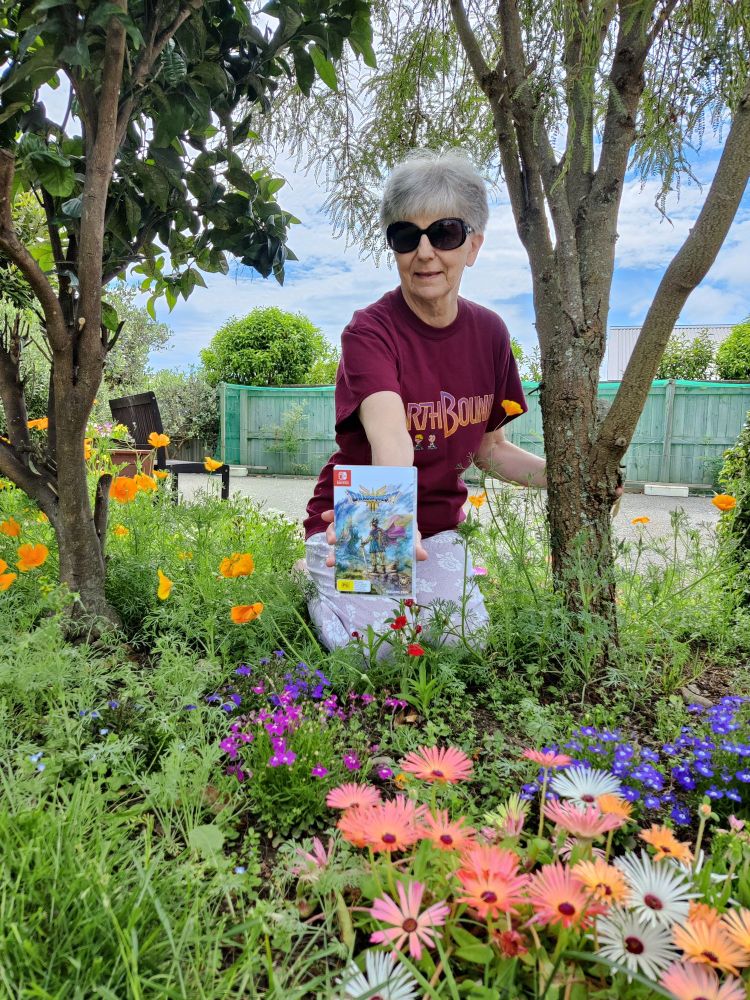 Me among colourful flowers in garden, between two trees, holding copy of DQ III, wearing Earthbound t-shirt.