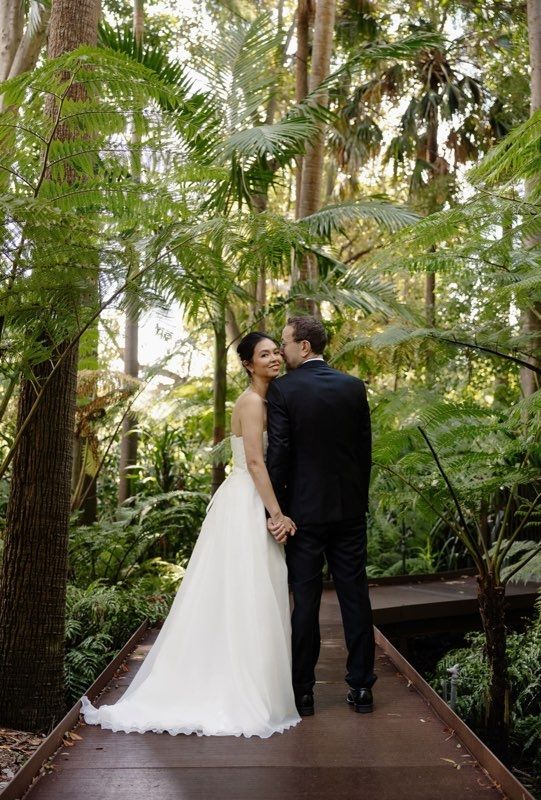 Just married couple, with husband's back facing the camera, looking sideways towards his bride. The bride facing sideways, looking over her shoulder towards the camera.