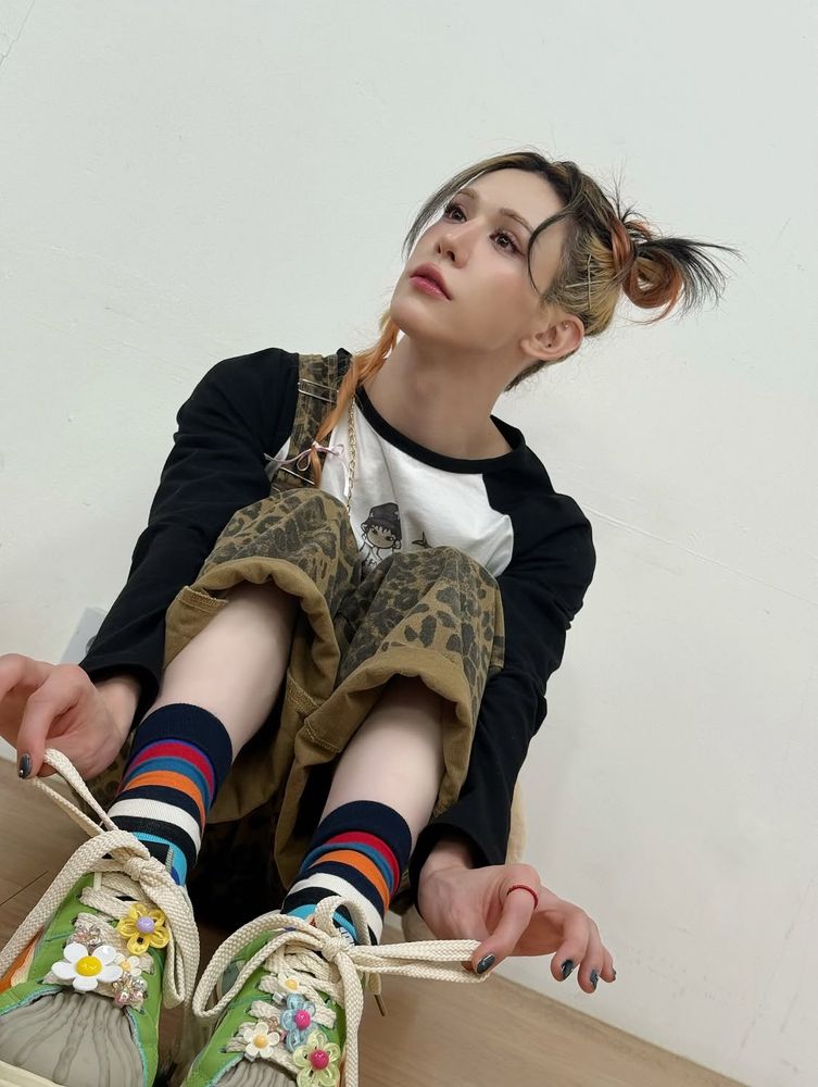 Wumuti sitting on the floor, legs up and knees cutely together, playing with his shoelaces. He’s wearing green sneakers with beaded flower charms on them, colorful striped socks, leopard-print overalls, and a baseball tee with a cartoon character on it. His hair is a variety of different colors (dark, bleached, orange) and mostly pulled back in a messy loop though one lock falls beside his face and is tied off with a little ribbon.