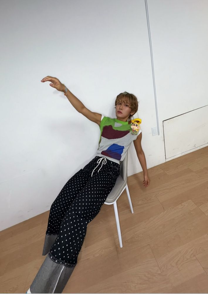 Hyun sitting in a chair and leaning back, one arm raised and leggies stuck out real far, the camera angle making them look impossibly long. His outfit is blessedly simple, I’m getting tired of typing. His trousers are black with little white polka dots and hugely long turn-up cuffs. He’s wearing a sleeveless shirt with abstract blocks of green, grey, matron, and blue. He has a charm of a cartoon character I know is recognizable but don’t know the name of on his shoulder. (Little kid with big eyes and yellow shirt.) His dark blond hair in styled in waves and long enough to cover his eyebrows and the tops of his ears. 