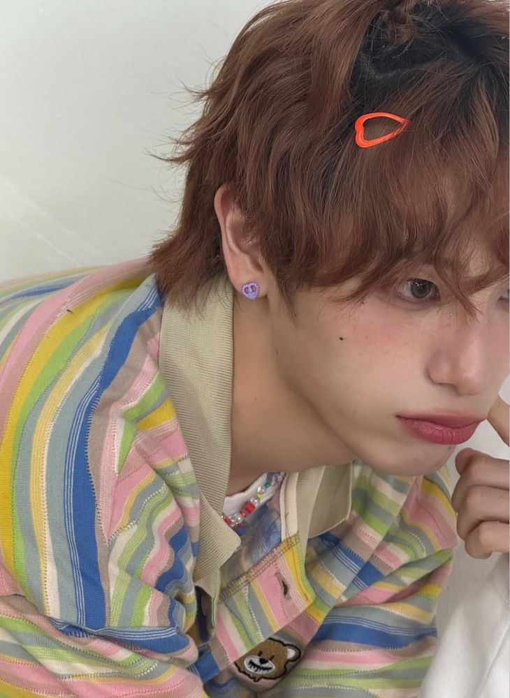 Haru seen only from mid-chest up, leaning forward until part of his face is out of frame to the right. He’s wearing a polo shirt with thin stripes of pastel pink, yellow, green, white, and darker blue, with a taupe collar and a little teddy bear face appliqué on the chest. A hint of a plastic beaded necklace can be seen. He has little plastic heart earrings and an orange heart clip in his slightly wavy mid-length red-brown hair.