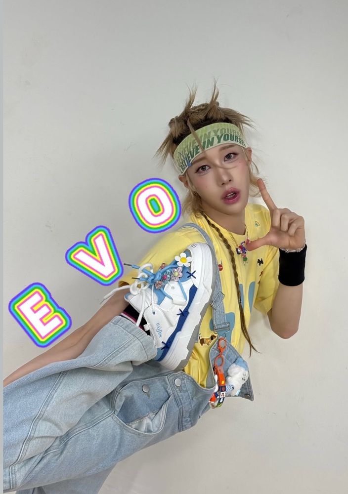 Rui standing up with his right leg pulled all the way back and up do his foot is at his armpit. The letters E V O have been added going up his back and his left hand is making a backwards L to finish the word he wears blue and white platform sneakers decorated with flower charms, light wash denim overalls buckled over only one shoulder and with charms hanging off, a yellow t-shirt, a beaded charm necklace, and a green headband that says “New York” over and over. His dark blond hair is in spiky messy knots on top of his head except for one long braided rattail.
