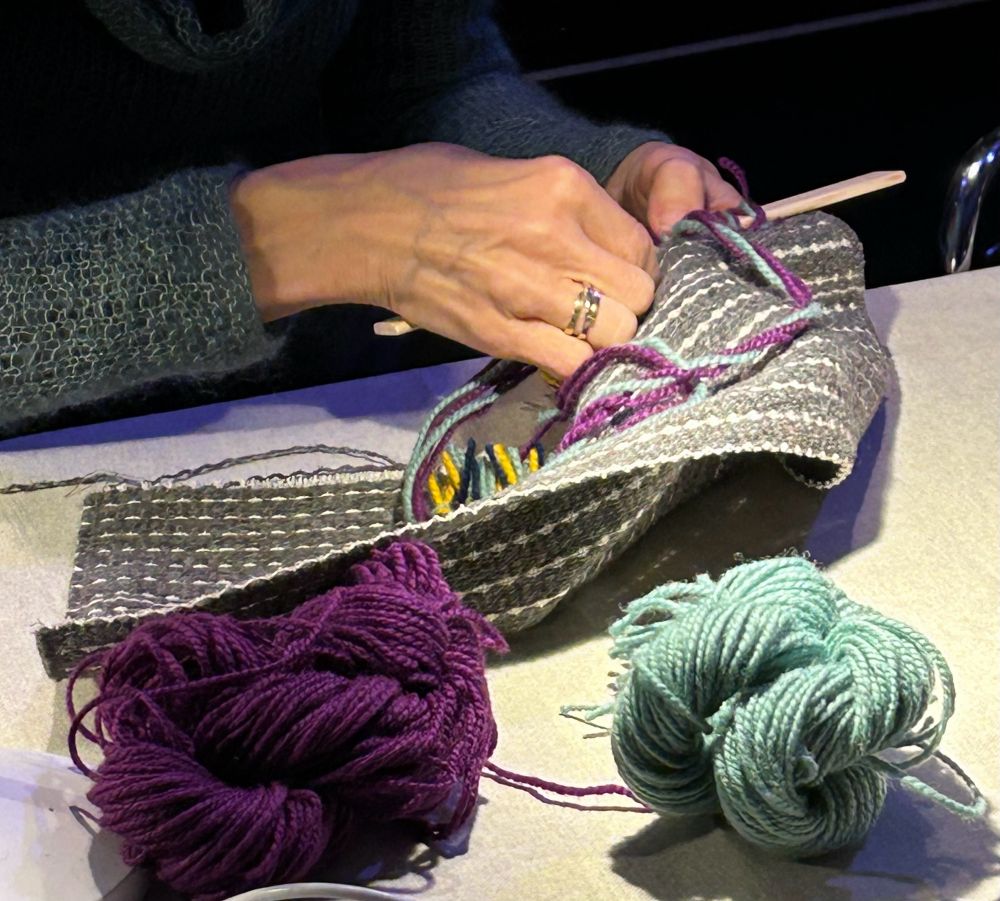 Hands creating a rya rug. Two balls of yarn in the foreground. 
