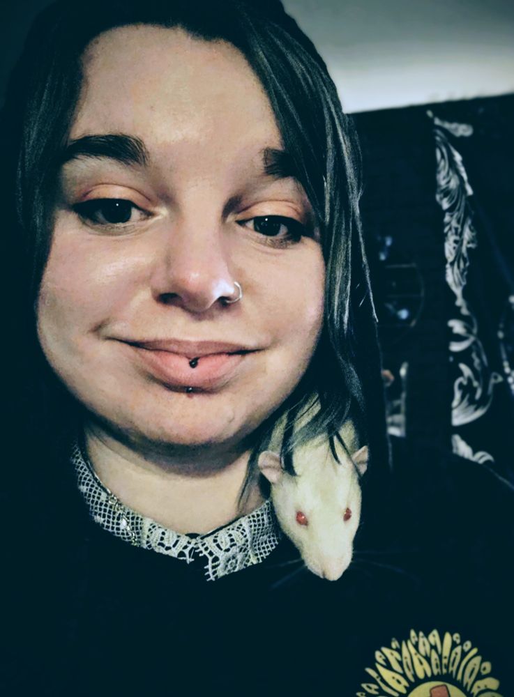 A portrait photo of a woman, with facial piercings and orange 'winged' eye makeup , looking directly into the camera smiling. She is wearing a black hoodie (hood is up) and a white collar, and a yellow sunflower logo is on the breast. A red-eyed cream and white (fawn) dumbo rat peaks his head out from her hood. Behind her is a black and white tapestry and a small crescent moon shelving unit. 