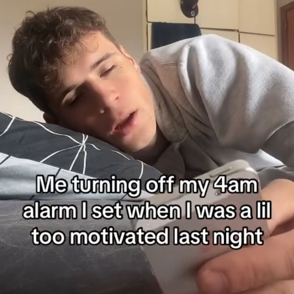 This meme shows a young man, looking tired and slightly annoyed, lying in bed with his eyes barely open. He is holding a smartphone in his hand as he appears to turn off an alarm. The caption reads: “Me turning off my 4am alarm I set when I was a lil too motivated last night.”