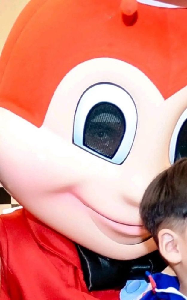 Close-up of a mascot costume with a large cartoonish red and white face. The mesh eye holes reveal a slightly visible human eye staring intently from within the costume. A child’s head is partially visible in the bottom right corner, wearing a blue outfit. The image has an unsettling vibe due to the visible human eye behind the cartoon face.