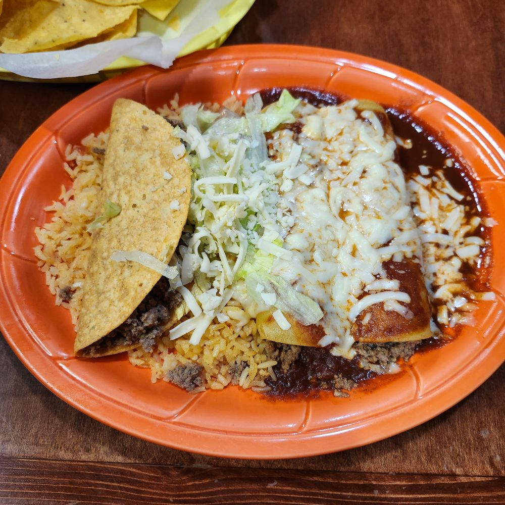 Plate at a mexican restaurant that has rice, one hard shell taco with ground beef, and two beef enchiladas smothered in red sauce and cheese. The taco toppings of lettuce and cheese are between everything
