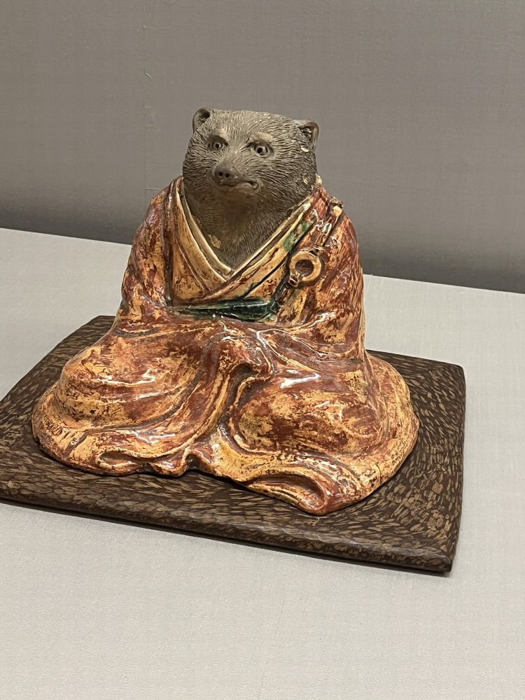 着物を着て正座するタヌキの陶器像。リアルな毛並みの顔と、赤茶色の艶のある衣が特徴で、木製の台座に載っている。