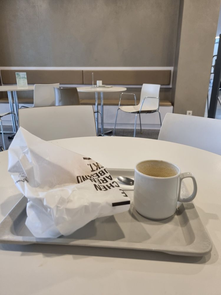 Tablett auf einem Tisch in einer Cafeteria, die Möbel und Wände sind in weiß und beige gehalten. Auf dem hellgrauen Tablett eine weiße Bäckertüte und eine weiße Tasse Kaffee.