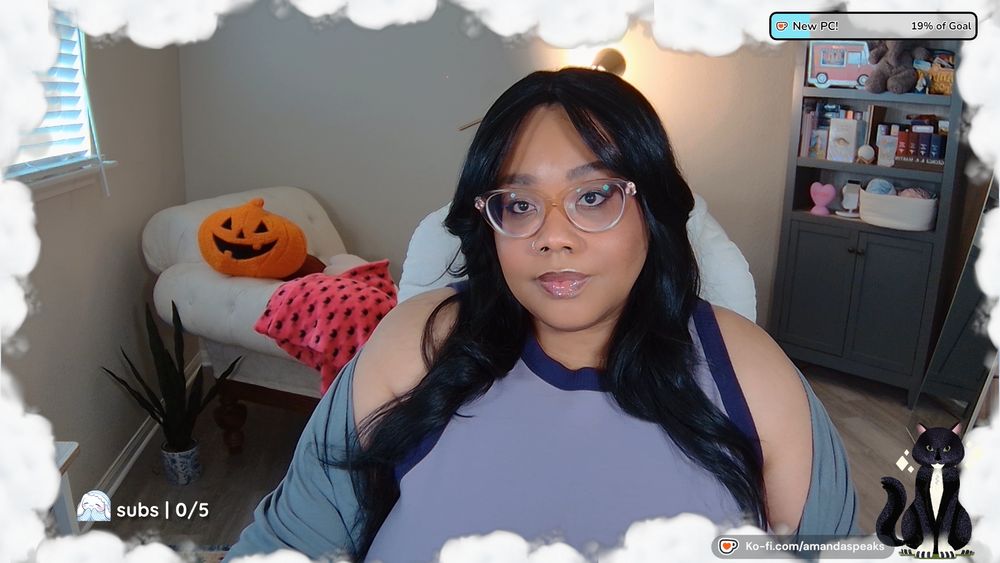 An image of Amanda; a Black person with long black hair and curtain bangs. They are wearing a light purple top with dark purple accents and a gray cardigan. Behind her is a cream colored chaise lounge with a jack-o-lantern plushie and hot pink throw blanket with a black bat pattern.