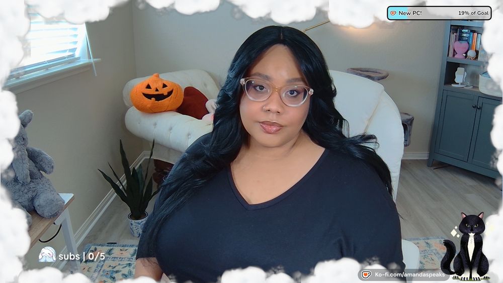 An image of Amanda; a Black person with long black hair and curtain bang. They are wearing a black v-neck top and sheer gold framed glasses.