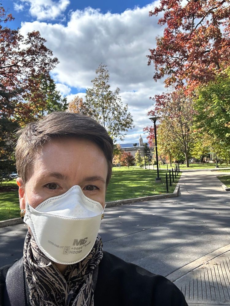 A white woman wearing a 3M mask, with autumn foliage and Penn State's Old Main campus behind her. 