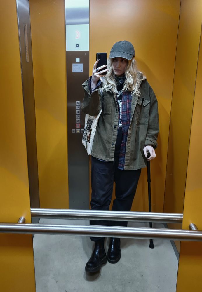 elevator selfie showing Newt wearing an oversized brown/tan corduroy jacket, black/burgundy plaid shirt, band tshirt and black cargo pants with doc marten boots and a khaki cap. he's lenaing on a cane and has a cream colored tote bag with patches on his shoulder. their blonde hair is loose and they look tired. 