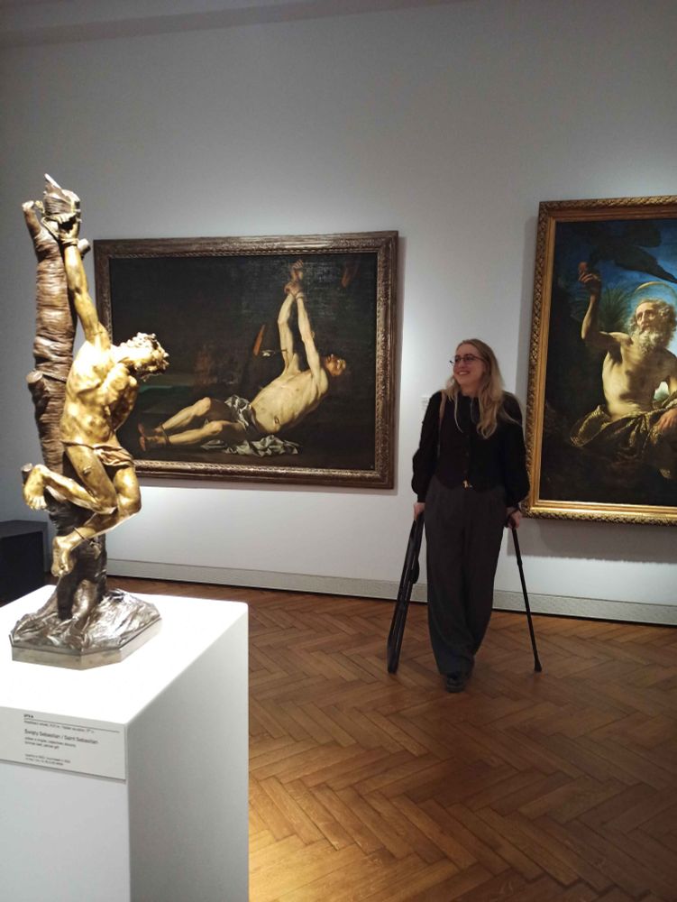 photo of newt, a white androgynous person dressed in all black with blonde hair standing in front of a large painting of a figure laying on the ground with his hands tied above and arrows piercing his side. there is a bronze statue of the same saint in the forgeround, tied to a tree and slumped to the side. newt is leaning on a cane in one hand and a fildable chair in the other and grinning at the view.
