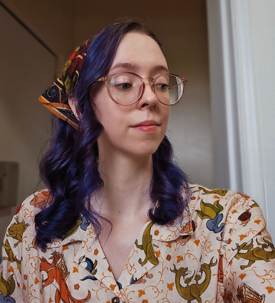 A selfie of a white nonbinary person with round translucent purple glasses and long, wavy purple hair, looking slightly to the right. They're wearing a bandana patterned with a stylized version of the unicorn tapestry, and a button up shirt with a pattern of medieval beasts and creatures 