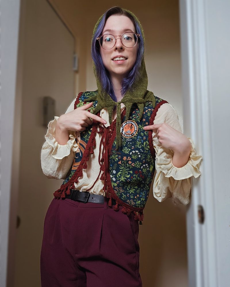 A white nonbinary person pointing to a sticker on their vest that says "Halloween Voter 2025" and has a trick-or-treating pigeon dressed as a witch in the center. They're wearing a Kermit the Frog medieval-inspired hood, a unicorn tapestry vest, a flowy linen shirt, and burgundy pants. 