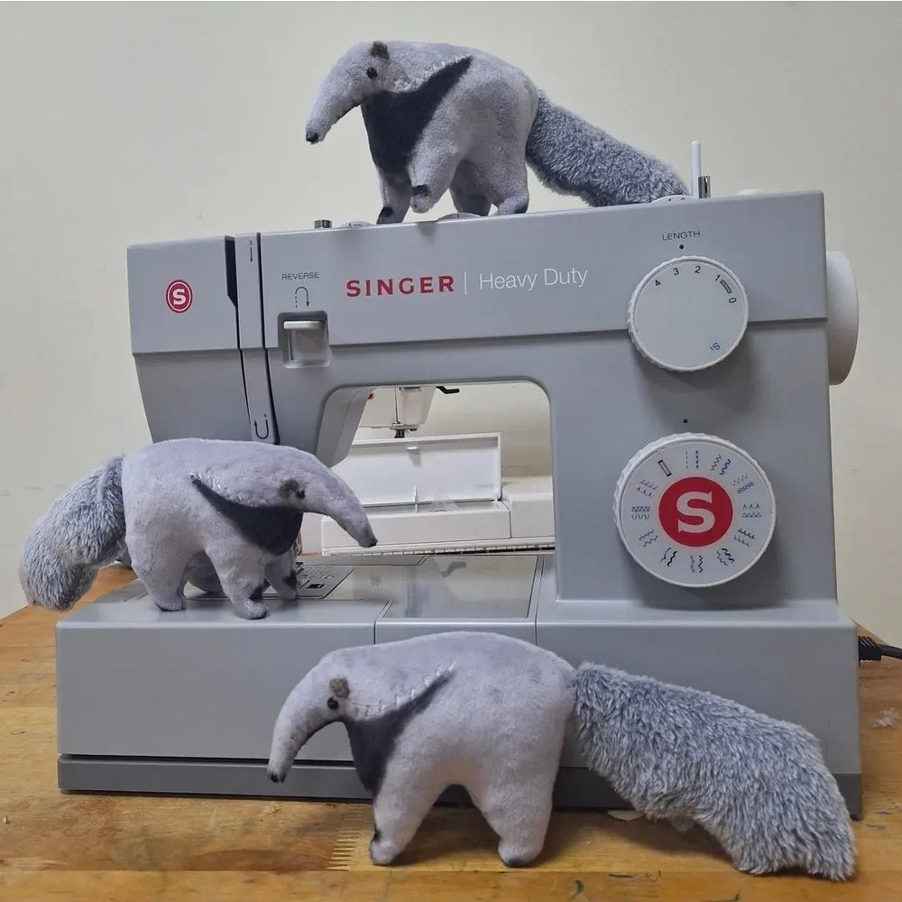 Three small giant anteater plushies sitting on and around a grey Singer sewing machine