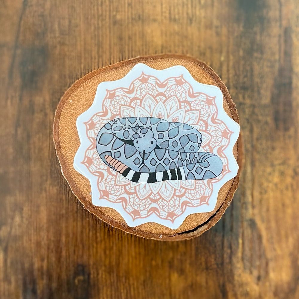 A waterproof vinyl sticker of a western diamondback with a tan, intricate mandala behind it. The sticker is sitting on a wood cutting with a wood background