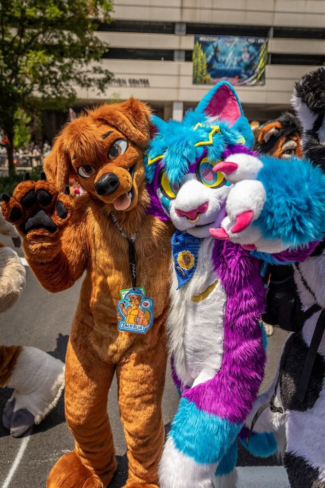 A brown dog fursuiter posing for a photo with a blue, purple and white car fursuiter