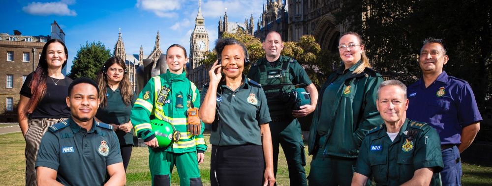 London Ambulance Service staff and volunteers in different uniform types, standing on a a sunny day with blue skies and Westminster Palace in the background.