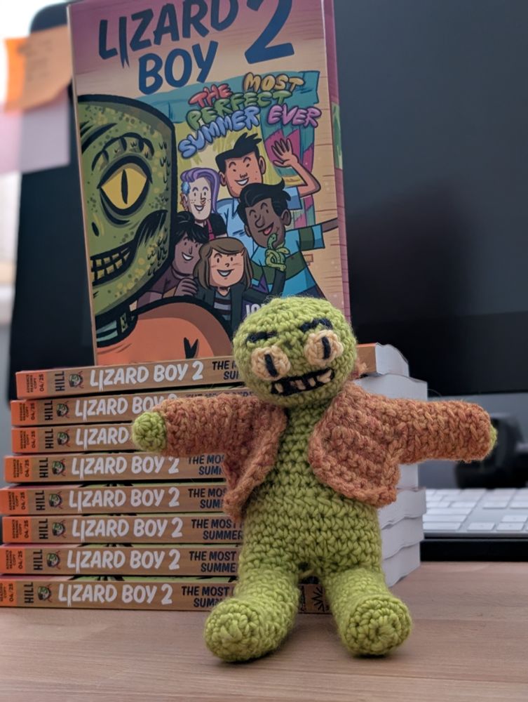 Lizard Boy 2 End of Summer Giveaway! Photo: a crocheted doll of Tommy the Lizard Boy in front of a stack of advanced reader copies of the middle grade graphic novel, Lizard Boy 2: The Most Perfect Summer Ever as well as a hard cover copy of the final book. 