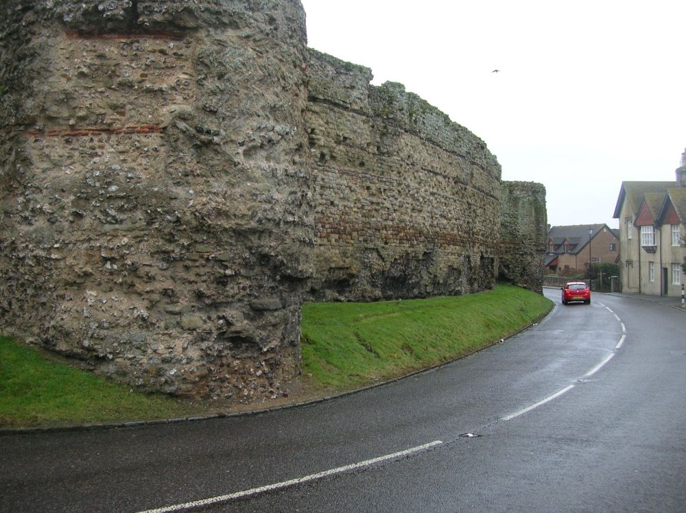 Towers of a huge stone and tile wall with a tarmac road and central white painted line in front. Red car and small modern buildings in the distance