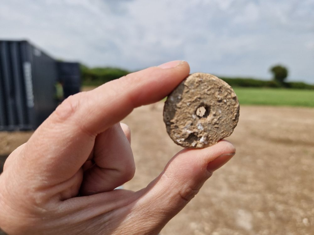 A small fragment of prehistoric pottery, reworked into a perforated disc, is held between the thumb and forefinger of an archaeologist with a ploughed field, hedge, sky and metal cabin in the background 