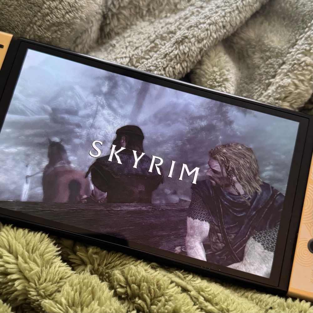 a switch laying on green blankets. the screen shows the opening screen from The Elder Scrolls V: Skyrim. one screen one man is being transported in a horse drawn cart by a soldier. 