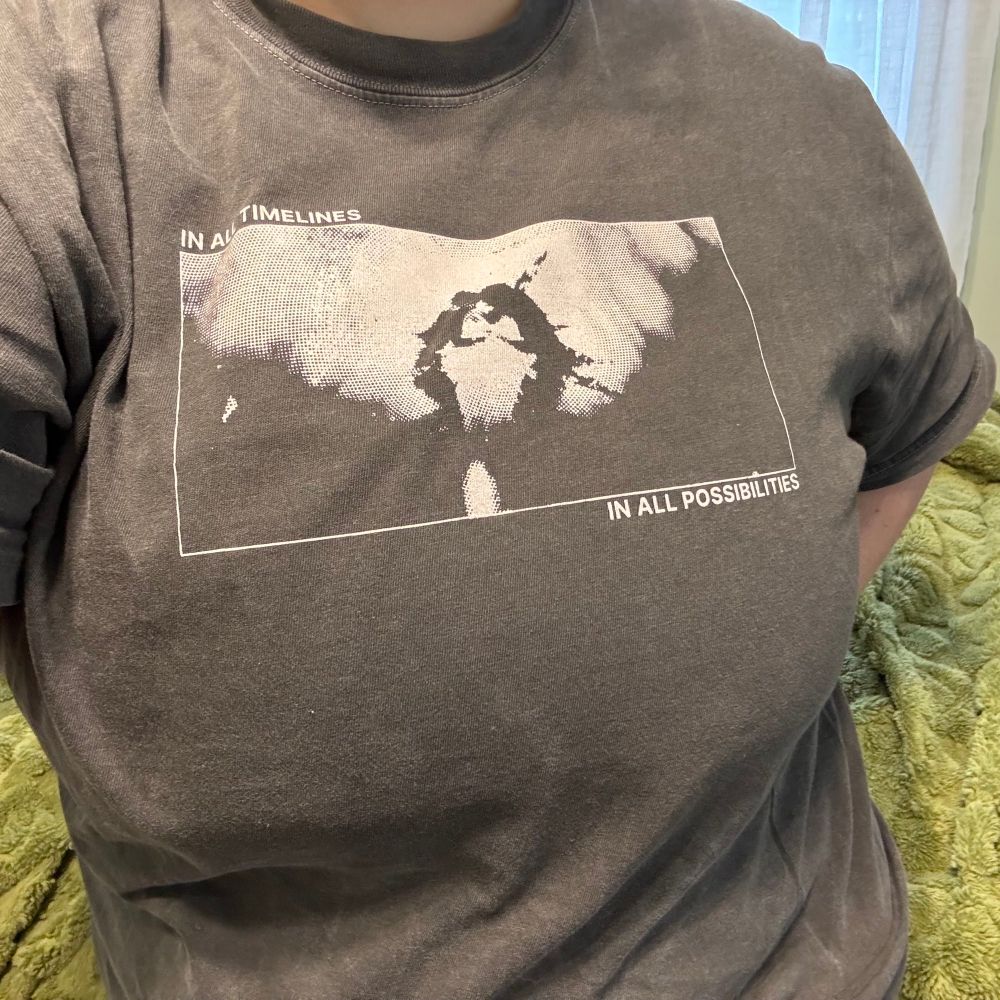 person wearing grey shirt. grey shirt has a Jayvik design from show arcane. It shows two characters with their heads bent together and the words “in all timelines/ in all possibilities 