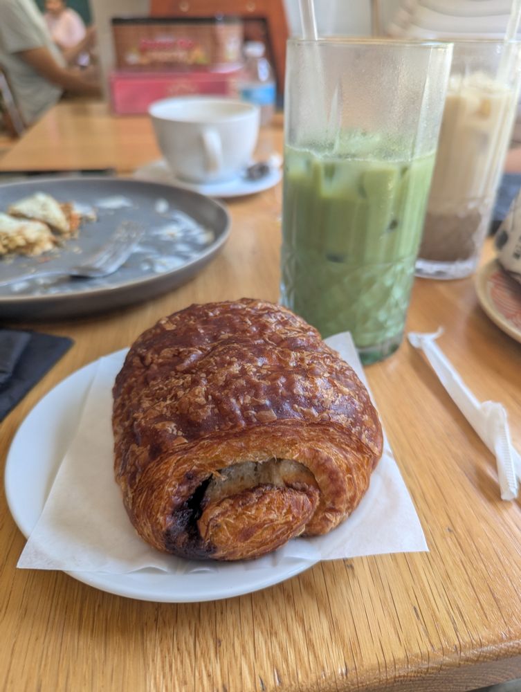 Quick cafe photo of a pain au chocolat and glass of matcha, with other coffees and teas and empty plates visible in the background